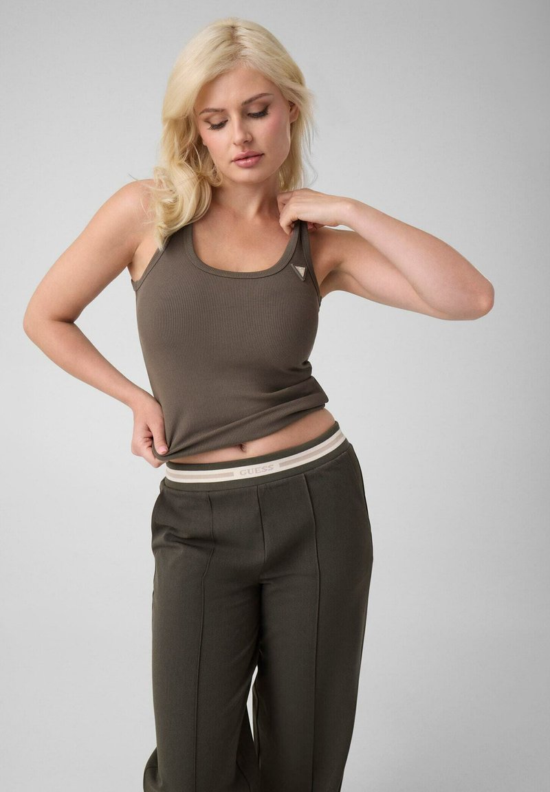 Femme blonde portant un débardeur olive et un pantalon ajuste son haut tout en regardant vers le bas sur un fond gris uni.