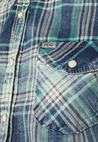Chemise en flanelle à carreaux bleu et vert avec un design classique, des boutons-pression et une poche poitrine ornée d'une étiquette logo. Texture douce.