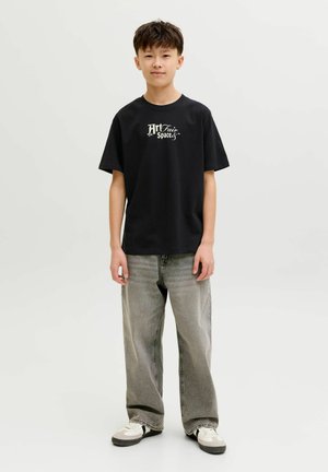 Nastolatek stojący w czarnej koszulce z napisem "Art Fair Spaces", luźnych szarych dżinsach i białych trampkach na jasnym, jednolitym tle.