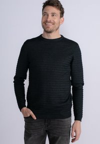 Maglione verde scuro con texture e righe orizzontali, collo a giro e maniche lunghe, abbinato a pantaloni in denim grigi.