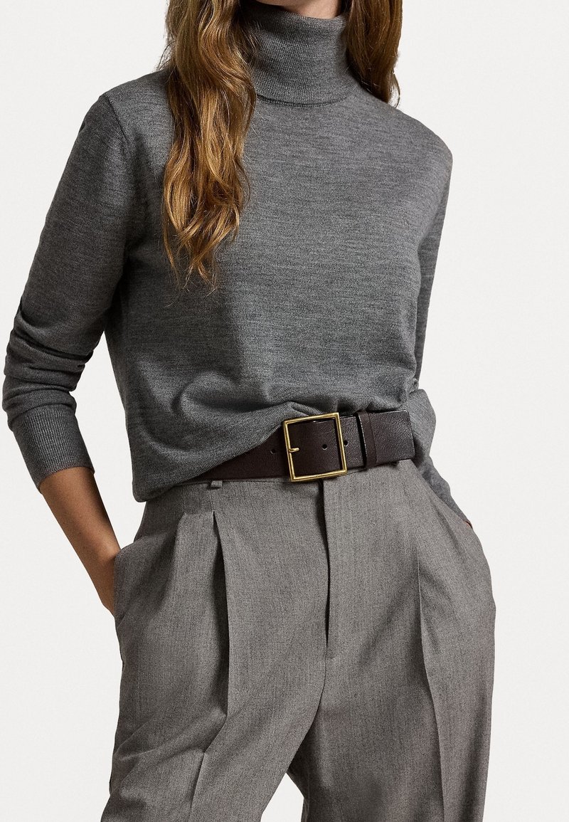 Femme portant un pull à col roulé gris rentré dans un pantalon plissé gris taille haute avec une ceinture marron foncé et une boucle dorée.