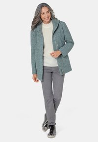 Türkise, strukturierte Jacke mit Kapuze, vorderem Druckknopfverschluss und zwei Seitentaschen, kombiniert mit einem cremfarbenen Pullover und grauen Hosen.