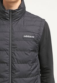 Svart vadderad väst med hög krage, med horisontell sömnad och dragkedjestängning. Inkluderar en rektangulär Adidas-logotyp på bröstet.