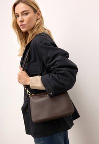 Brązowa skórzana torba na ramię z teksturowaną powierzchnią, zamknięciem na zamek i złotymi akcentami, noszona przez modelkę w ciemnej kurtce i beżowym swetrze.