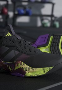Chaussure de sport noire avec des détails de semelle jaune néon, violet et à motif, reposant sur un sol de salle de sport sombre avec des poids flous en arrière-plan.