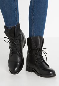 Botines de cuero negro con cordones, punta redondeada y tacón bajo. Detalles de puños doblados y textura suave. Llevados con jeans de mezclilla azul.