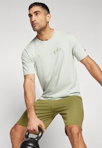 Hellgrünes Baumwoll-T-Shirt mit gesticktem "Champion"-Logo und olivgrünen Shorts. Das Modell hält eine schwarze Kettlebell. Glatte Textur.