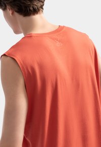 T-shirt orange sans manches en tissu en coton doux, avec un petit logo de ciseaux imprimé sur le dos près de l'encolure.