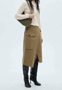 Falda midi caqui con bolsillos laterales, combinada con un suéter de punto trenzado color crema y botas altas negras. Un bolso verde olivo completa el look.