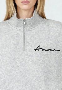 Sweat-shirt gris à fermeture éclair avec un col montant, en tissu texturé et broderie noire « Amour » sur la poitrine.
