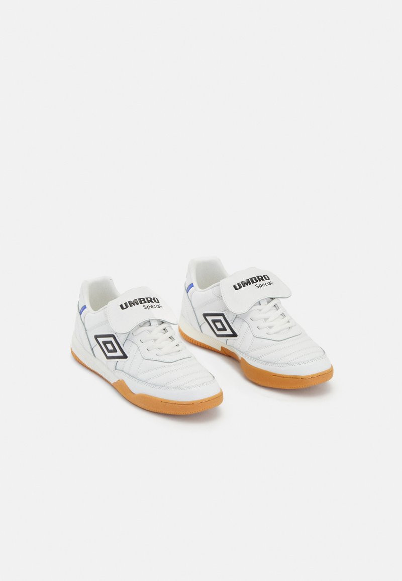 White athletic shoes with a rubber gum sole, featuring a black and white logo, lace-up front, and velcro strap for added support.