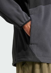 Pull en polaire gris foncé avec une surface texturée, présentant un panneau contrastant, des poignets élastiques et une poche latérale.