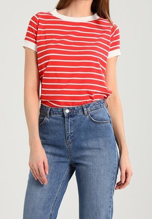 Kvinde iført en rød og hvid stribet kortærmet skjorte stoppet ned i højtaljede blå jeans, stående med armene afslappede.