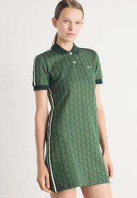 Rochie polo verde cu un model geometric, cu mâneci scurte și guler. Prezintă dungi laterale albe în contrast și un mic logo pe piept.