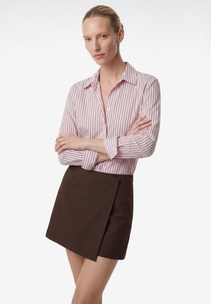 Femme aux cheveux blonds courts portant une chemise à rayures roses et blanches à boutons et une jupe mini asymétrique marron foncé, debout les bras croisés.