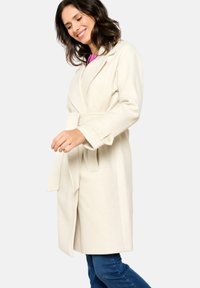 Cream wool coat with a wrap design, wide lapels, and long sleeves. Features a tie belt at the waist and is knee-length.