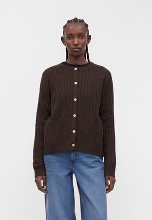 Vrouw met gevlochten haar die een donkerbruine gebreide cardigan met knopen en lichtblauwe jeans draagt, staand tegen een effen witte achtergrond.