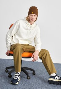 Crème hoodie met logo, kaki broek, bruin beanie en marineblauwe sneakers. Hij zit op een oranje bureaustoel tegen een grijze achtergrond.