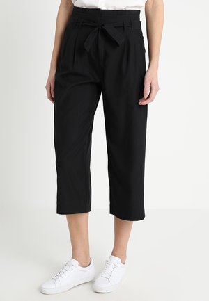 Pantaloni neri a vita alta, realizzati in un tessuto leggero, con cintura da annodare, gamba plissettata e tasche laterali. Abbinati a sneakers bianche.