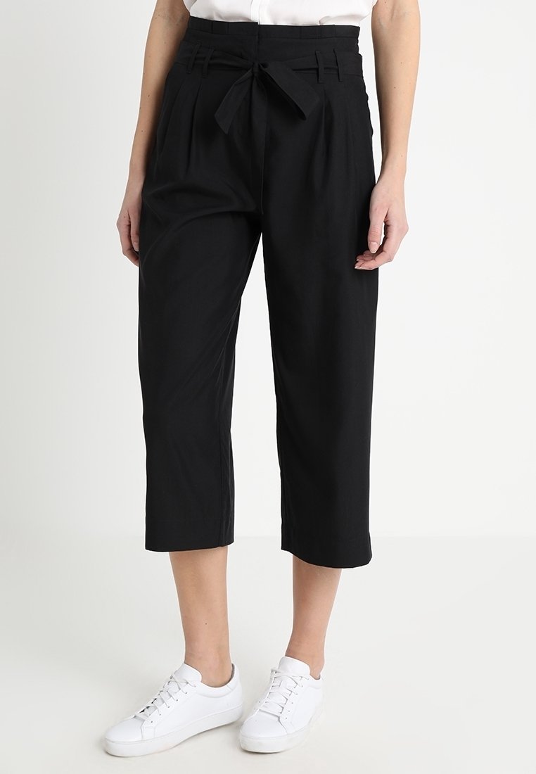 Zwarte cropped broeken gemaakt van een lichte stof, met een hoge taille en een strikceintuur, geplooide voorkant en zijzakken. Gecombineerd met witte sneakers.