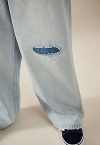 Lyseblå denimjeans med en markant rift med blå lapning, let frynsete kanter og en afslappet pasform, kombineret med sorte sneakers.