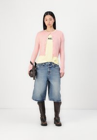 Cárdigan corto de color rosa claro con botones en la parte delantera, camiseta amarilla con texto negro, pantalones cortos de mezclilla azules holgados y botas marrones hasta la rodilla.