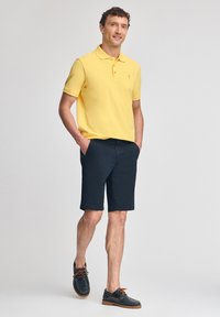 Gul polotröja, marinblå shorts och bruna seglarskor med orange snören. Outfiten har en klassisk passform och en avslappnad design.