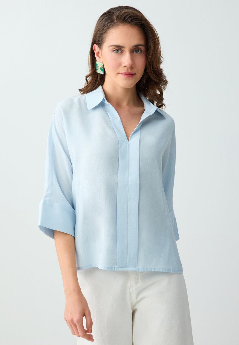 Femme aux cheveux bruns ondulés porte un chemisier bleu clair oversize, de grandes boucles d’oreilles géométriques vertes et dorées, ainsi qu’un pantalon blanc sur un fond uni.