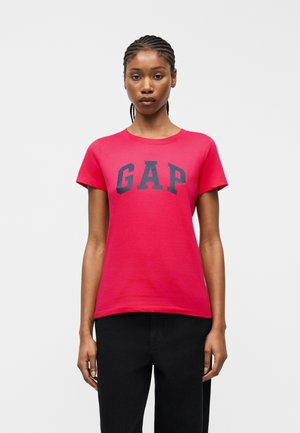 Fonott hajjal rendelkező nő élénkpiros "GAP" logós, sötétkék betűkkel díszített pólóban és fekete nadrágban, egyszerű fehér háttér előtt áll.
