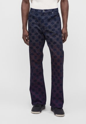 Personne portant un pantalon bleu foncé avec un motif géométrique violet, des chaussures noires et un haut blanc texturé, debout devant un fond neutre.