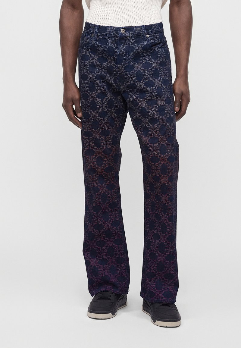 Personne portant un pantalon bleu foncé avec un motif géométrique violet, des chaussures noires et un haut blanc texturé, debout devant un fond neutre.