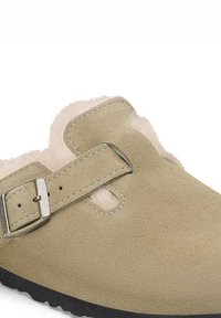 Zapato deslizante de ante beige con una superficie texturizada, correa ajustable con hebilla y un suave forro de piel de pelo visible en la apertura y los bordes.