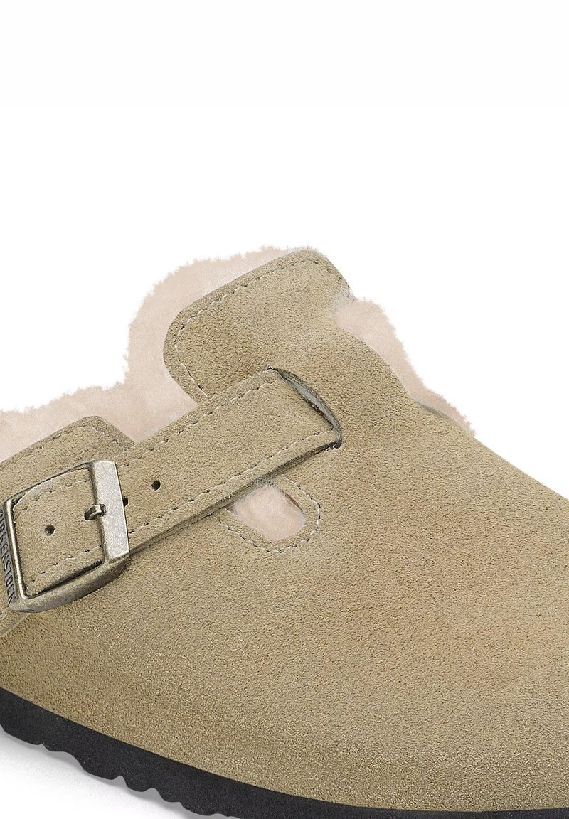 Chaussure slip-on en suede beige avec une surface texturée, une sangle à boucle ajustable et une douce doublure en fourrure visible à l'ouverture et sur les bords.