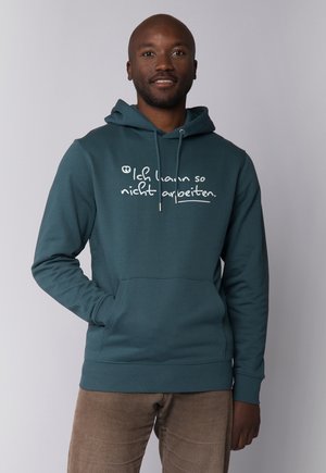 Man in een teal hoodie met Duitse tekst "Ich kann so nicht arbeiten" en bruine corduroy broek, staand tegen een effen lichte achtergrond.