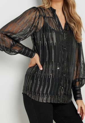 Femme portant un chemisier noir boutonné avec des manches longues transparentes et des rayures verticales de sequins argentés, assorti à un pantalon noir.