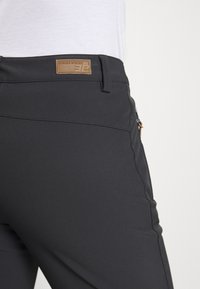 Primer plano de la cinturilla de unos pantalones negros con un parche de logo marrón y un bolsillo con cremallera en el lado derecho, llevados con una parte superior blanca.