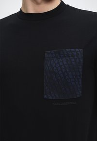 Άνδρας με μαύρο μπλουζάκι με στρογγυλή λαιμόκοψη και σκούρο μπλε τσεπάκι στο στήθος με μοτίβο επαναλαμβανόμενου κειμένου και διακριτικό λογότυπο "KARL LAGERFELD" από κάτω.