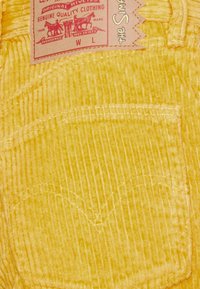 Žlutá kordová tkanina má vertikální žebrování, nášivku s logem značky a kapsu se švy.