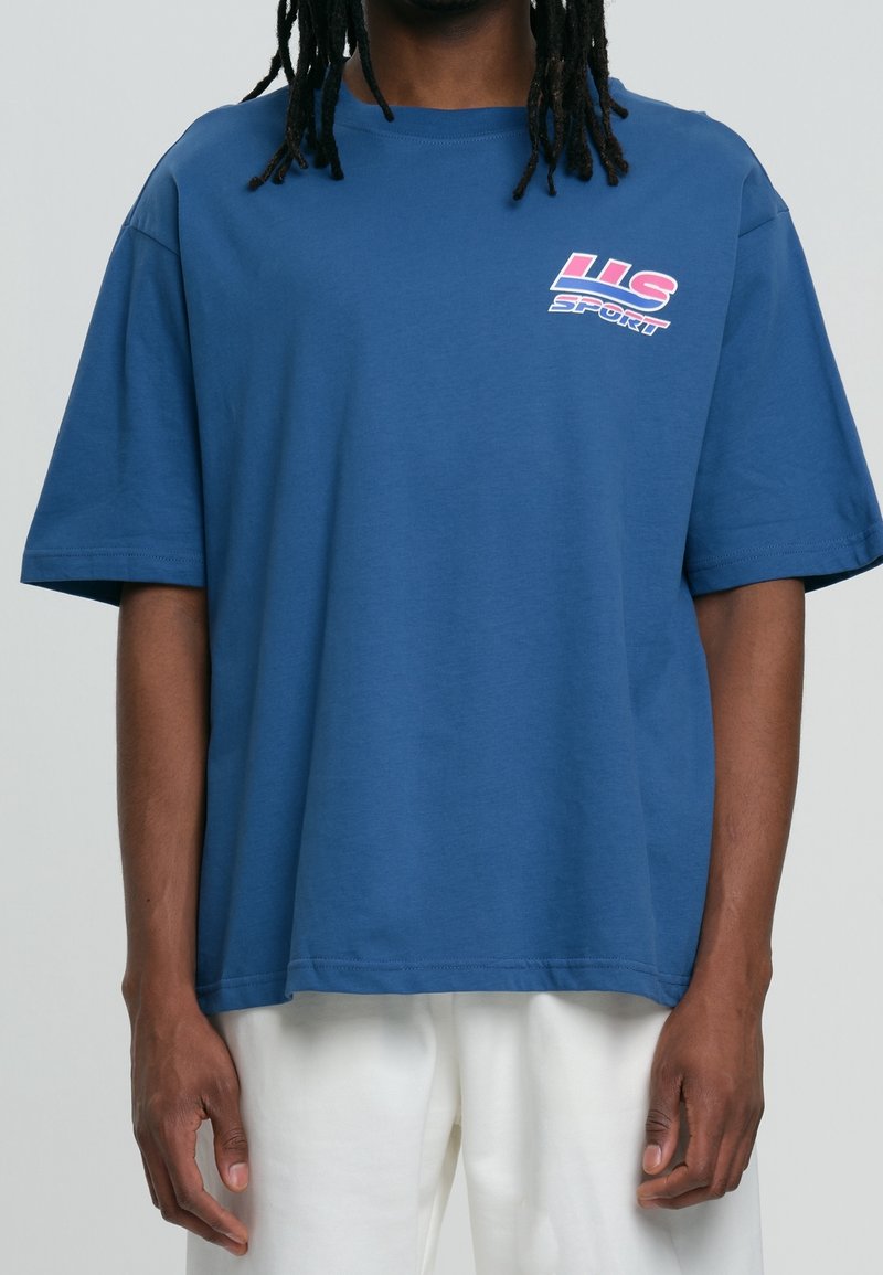 Personne portant un t-shirt bleu ample avec un logo rose et blanc « US Sport » sur le haut gauche de la poitrine, associé à un pantalon blanc.