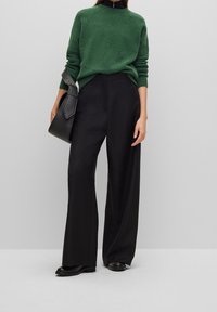 Green knit sweater with a crew neckline, paired with high-waisted black wide-leg trousers. Model holds a black shoulder bag.