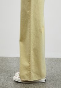 Pantaloni a gamba larga in cotone morbido giallo chiaro con finitura testurizzata, caratterizzati da un dettagli al lato e che coprono sneakers bianche con accenti rossi.