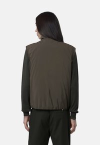 Gilet smanicato, di un marrone scuro, imbottito e dalla superficie liscia, con orlo elastico e collo alto. Indossato sopra una maglietta scura a maniche lunghe.