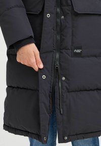 Svart polstret jakke med glidelås og trykknappfeste. Har et logo merke og sidelommer. Glatt tekstur med quiltet design.