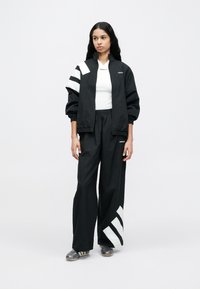 Svart atletisk jakke med hvite diagonale striper; matchende svarte bukser med hvite detaljer. Begge laget av lett, teksturert materiale.