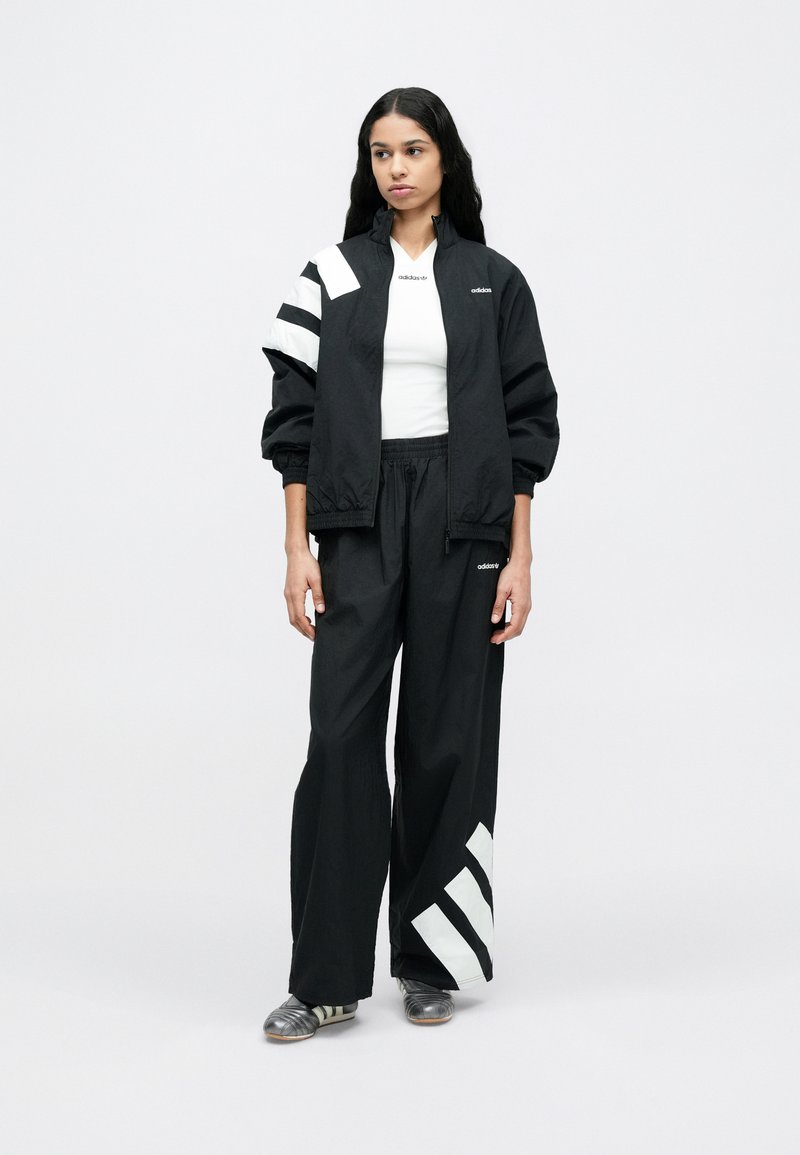 Sort atletikjakke med hvide diagonale striber; matchende sorte bukser med hvide detaljer. Begge lavet af let, tekstureret materiale.