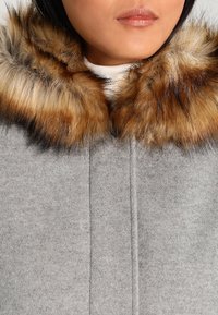 Grå kappe med blød tekstur, der har en pelskrave i nuancer af brun og beige. Designet inkluderer en lukning foran og er uden dekorationer.