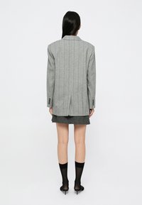 Une femme aux longs cheveux noirs se tient dos à nous, portant un blazer gris à rayures, une jupe sombre, des collants noirs transparents et des sandales à talons noires.