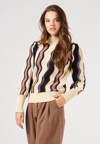 Jersey de punto con cuello alto, con patrones ondulados en crema, azul marino y marrón. Las mangas abullonadas y el dobladillo acanalado añaden textura. Se muestran pantalones marrones.