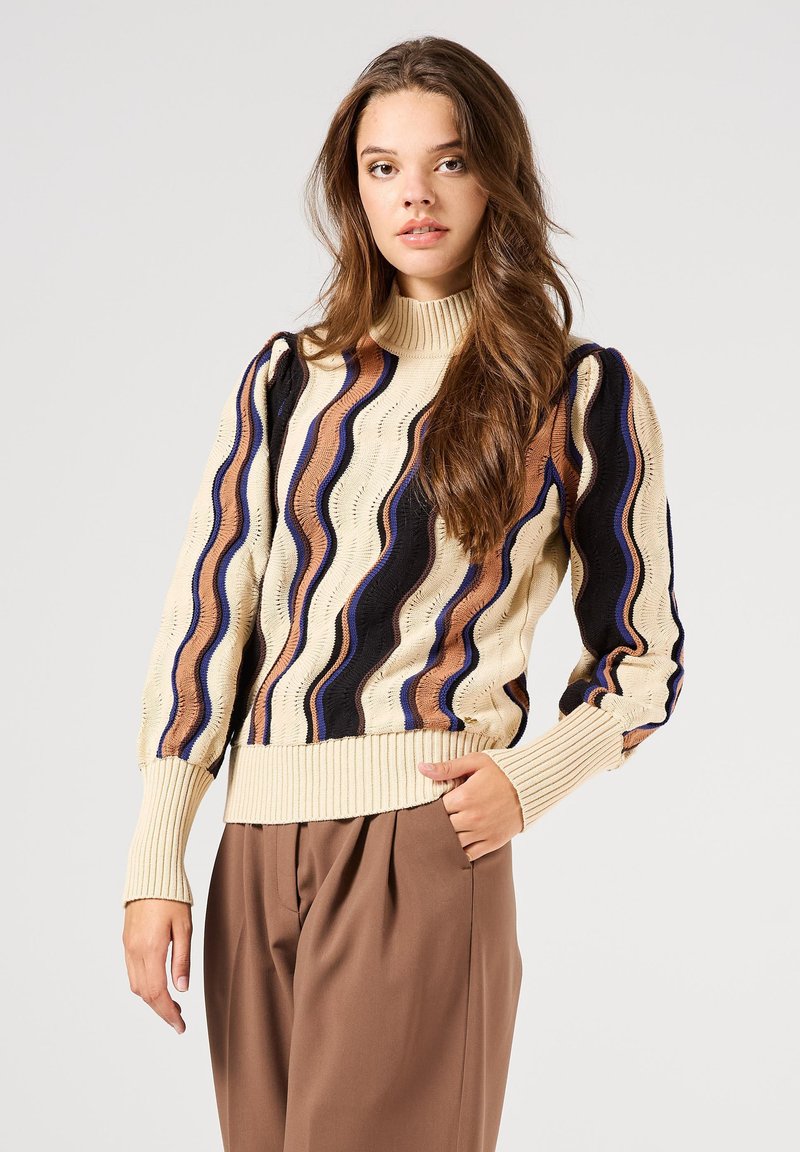 Jersey de punto con cuello alto, con patrones ondulados en crema, azul marino y marrón. Las mangas abullonadas y el dobladillo acanalado añaden textura. Se muestran pantalones marrones.
