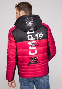 Chaqueta acolchada roja y negra con capucha, que presenta impresiones de texto, textura acolchada, bolsillo con cremallera y mangas en contraste.